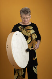 Middle aged woman with blonde hair looking down and playing a large white frame drum