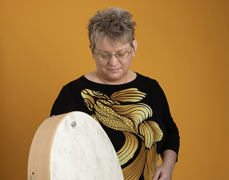 Middle aged woman with blonde hair looking down and playing a large white frame drum