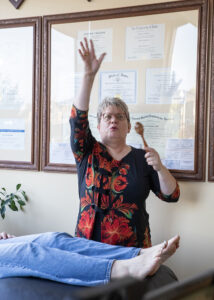 Kerri depicting core shamanic healing - Mammoth Hills Coaching and Wellness, LLC A middle aged woman with a rattle and reaching for sky above a woman lying on a massage table