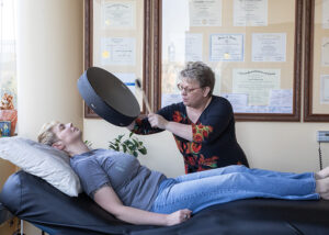 Kerri depicting shamanic drumming - Mammoth Hills Coaching and Wellness, LLC A middle aged woman drumming over another younger woman lying on a massage table face up.