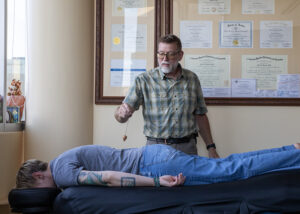 A middle-aged man assessing the spine of woman lying down on a massage table and wearing jeans and a t-shirt. He is using a pendulum to assess the energy field