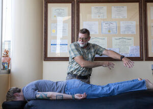Middle-aged man engaged in field repatterning over a woman lying face down on a massage table. He is wearing glasses and a short sleeved shirt and khakis and she is wearing a gray t-shirt and jeans