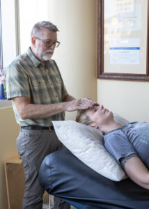 Todd providing another step of glymphatic draining a healing touch technique - Mammoth Hills Coaching and Wellness, LLC A middle aged man providing healing touch over the head of a woman lying down on a massage table face up.