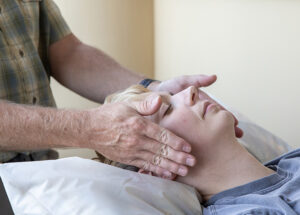 A man has his hands near the sides of the head of a resting woman on a massage table administering Healing Touch