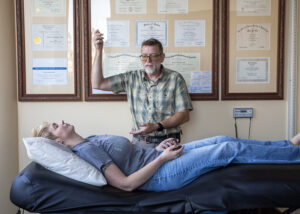 Todd providing fifth level Healing Touch - Mammoth Hills Coaching and Wellness, LLC Healthcare professional (man) performing a treatment on a patient lying on an examination table in a certificate-filled clinic room behind thems.