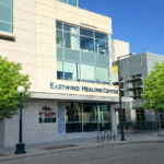 Exterior of Eastwind Healing Center from Linn Street. There is a side entrance on Linn Street and on College Street around the cornder. The Eastwind name is on the 2nd floor above Formosa restaurant, and there are green trees on either side.