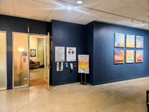 USE Eastwind Condo Entrance 1 - Mammoth Hills Coaching and Wellness, LLC Interior hallway of a healing center with dark blue walls, framed posters on the right, and an open doorway to a warmly lit waiting area on the left.