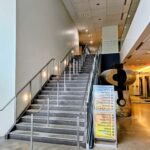 Indoor modern lobby with wide metal staircase and glass railing, right-side abstract stone sculpture, and a signboard listing events or speakers nearby