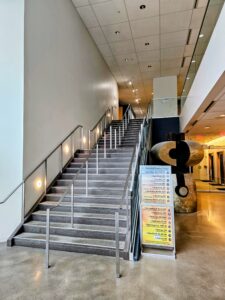 USE stairs at an angle 1 - Mammoth Hills Coaching and Wellness, LLC Indoor modern lobby with wide metal staircase and glass railing, right-side abstract stone sculpture, and a signboard listing events or speakers nearby
