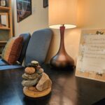 A stone cairn stack on a wooden base sits on a dark table beside a blue chair and a tall brown table lamp.