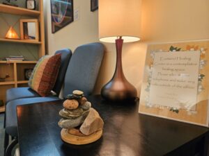 USE waiting room cairn 1 - Mammoth Hills Coaching and Wellness, LLC A stone cairn stack on a wooden base sits on a dark table beside a blue chair and a tall brown table lamp.
