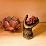 Bronze ornate vase with an orange floral arrangement beside a pink lotus sculpture on a wooden shelf under warm light.