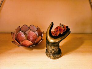 USE waiting room candle and brass hand 2 - Mammoth Hills Coaching and Wellness, LLC Bronze ornate vase with an orange floral arrangement beside a pink lotus sculpture on a wooden shelf under warm light.