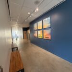 Long hallway with a blue accent wall displaying six framed sunset photos under track lighting and wooden benches along the white wall on the left.