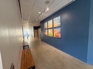 entrance hallway - Mammoth Hills Coaching and Wellness, LLC Long hallway with a blue accent wall displaying six framed sunset photos under track lighting and wooden benches along the white wall on the left.